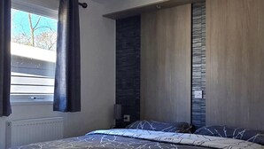 A modern bedroom with a large bed, a window with curtains, a built-in bench, and a wall-mounted light fixture. - Vakantiepark Kijkduin - 838 (Den Haag)