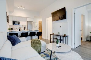 A modern living room with a sofa, a coffee table, and a dining area with chairs and a kitchen in the background. - Stylish Downtown Suite With Patio City Views (Kitchener)