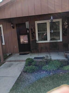 A house with a front door, two windows, a porch with chairs, and a small garden area.