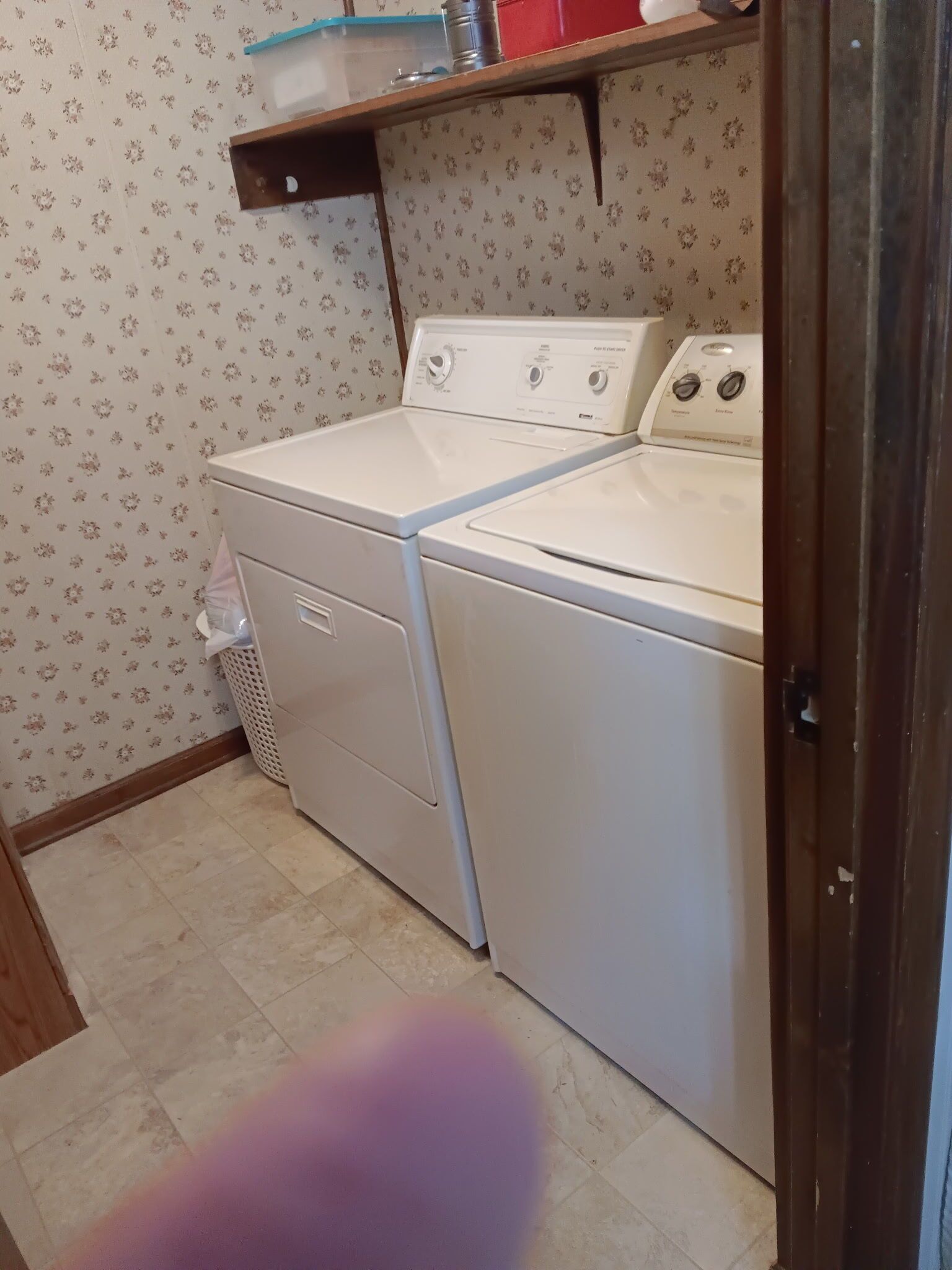 A laundry room with two stacked washers and dryers, a small shelf above, and a trash bin.