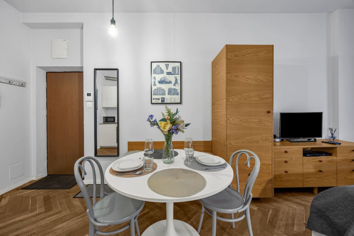 Quarto moderno com mesa de jantar redonda, duas cadeiras, um vaso com flores, televisão de tela plana e um armário de madeira.