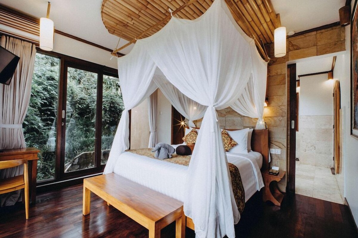 A room with a bed under a canopy, a bench, a bedside table, and a view of greenery outside.