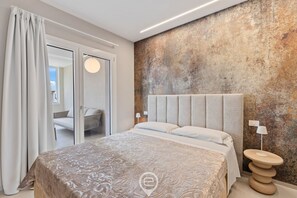 A modern bedroom with a large bed, a textured wall, a round light fixture, and a view of another room. - Appartamento Ginevra 3 (Arzachena)