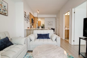 A living room with a sofa, a coffee table, and a TV stand. There is a kitchen in the background with wooden cabinets and a refrigerator. - Modern High-rise Condo w City Views Balcony (Kitchener)