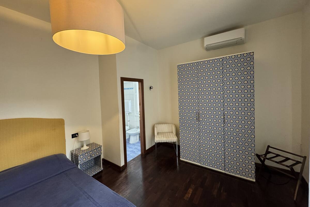 A bedroom with a bed, a bedside table, a lamp, a chair, a patterned wall, and a door leading to a bathroom.