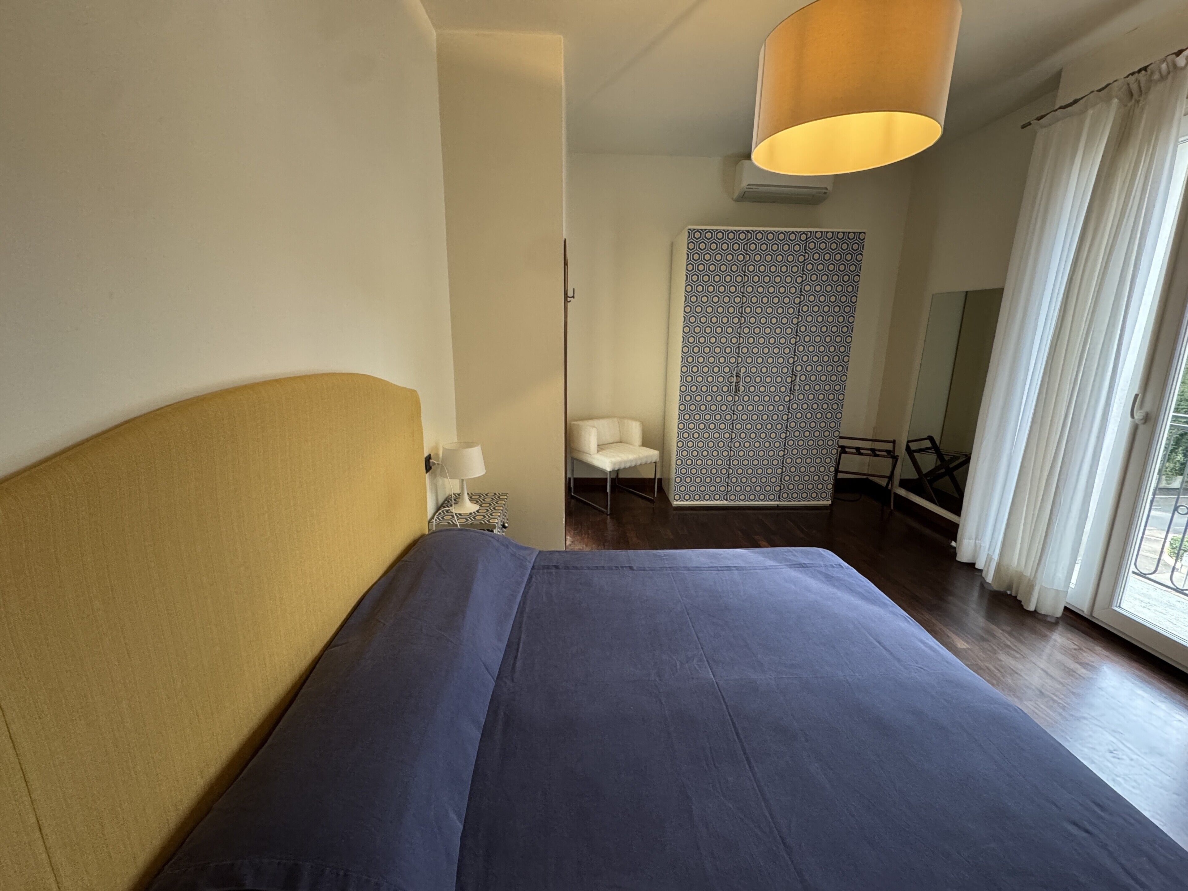 Ein Hotelzimmer mit Bett, Sessel, Wand mit Muster und Fenster mit Vorhängen.