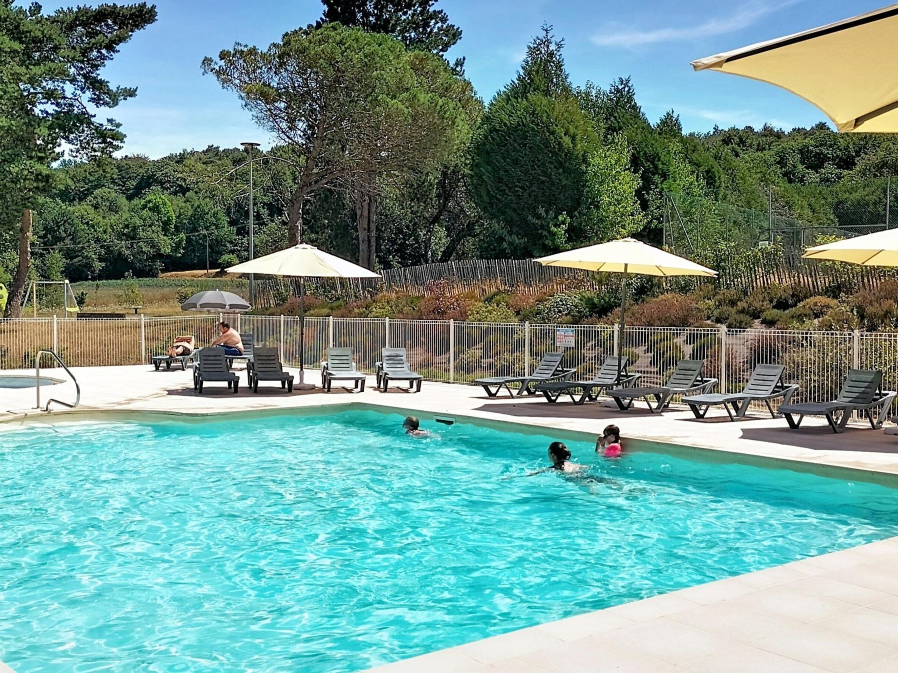 Una piscina con sillones de descanso y sombrillas, rodeada de una valla y árboles.