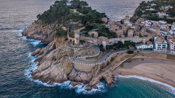 Vista aérea de uma vila costeira com uma estrutura fortificada, uma praia e um litoral rochoso.