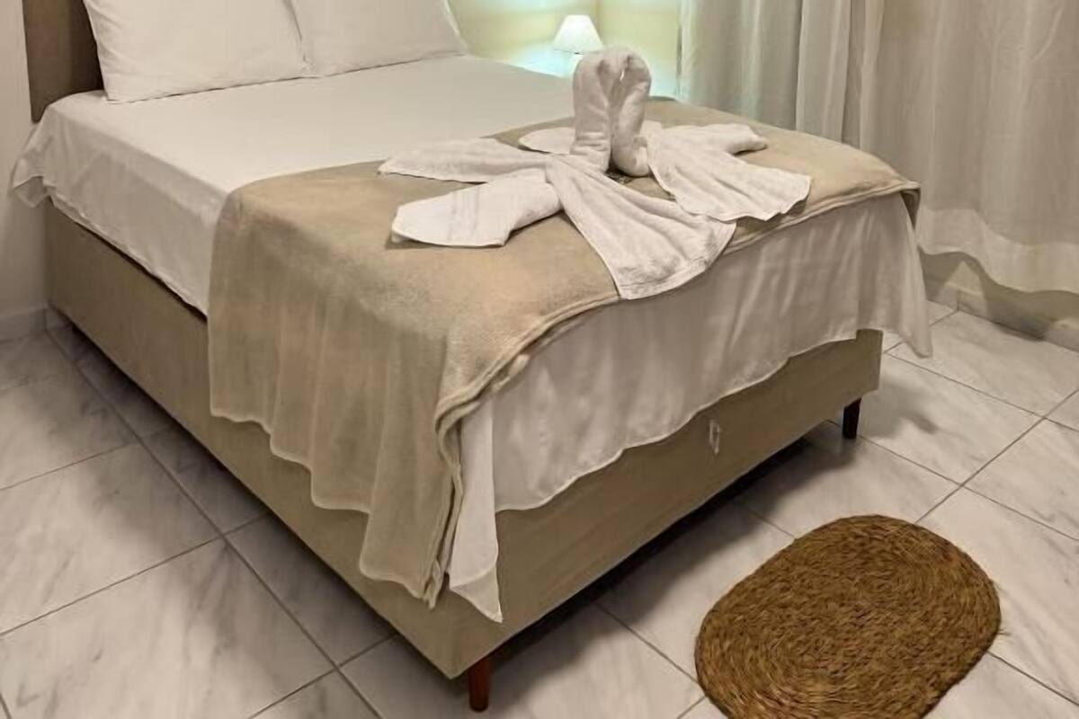 Una cama bien hecha con sábanas blancas y una toalla, una lámpara de noche y un tapete tejido sobre un suelo de baldosas.