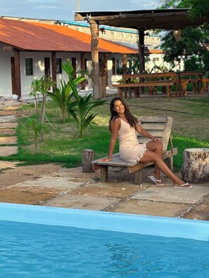 A person sitting on a wooden bench by a swimming pool with a building in the background. - Pousada Cajueiro Do Sol (Barreirinhas)