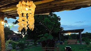 An outdoor dining area with a wooden table, a hanging light fixture, and a view of the surrounding garden and trees. - Pousada Cajueiro Do Sol (Barreirinhas)
