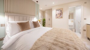 A modern bedroom with a large bed, a headboard with vertical lines, a painting on the wall, and a bathroom visible through an open door. - 108 Shirland Road (London)