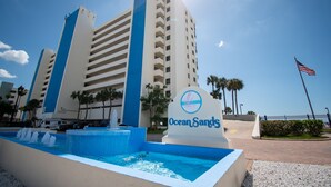 Un hotel con un letrero que dice 'Ocean Sands', un edificio grande de varios pisos, una piscina con fuentes de agua y una bandera estadounidense.