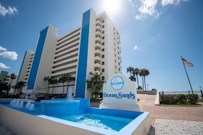 A hotel with a sign reading 'Ocean Sands,' a large building with multiple floors, a pool with water fountains, and an American flag. - ☀Stunning Views🐬Beachfront Condo🌴Ocean Sands II-602🐠Heated Pool/Hot Tub/Grills (Madeira Beach)