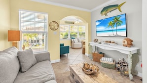 A living room with a sofa, a coffee table, a TV, and a wall-mounted shelf with decorative items. - 🏖️2 Min Walk to Beach🌊Prime Location☀Barefoot Beach Resort E206 (Indian Shores)