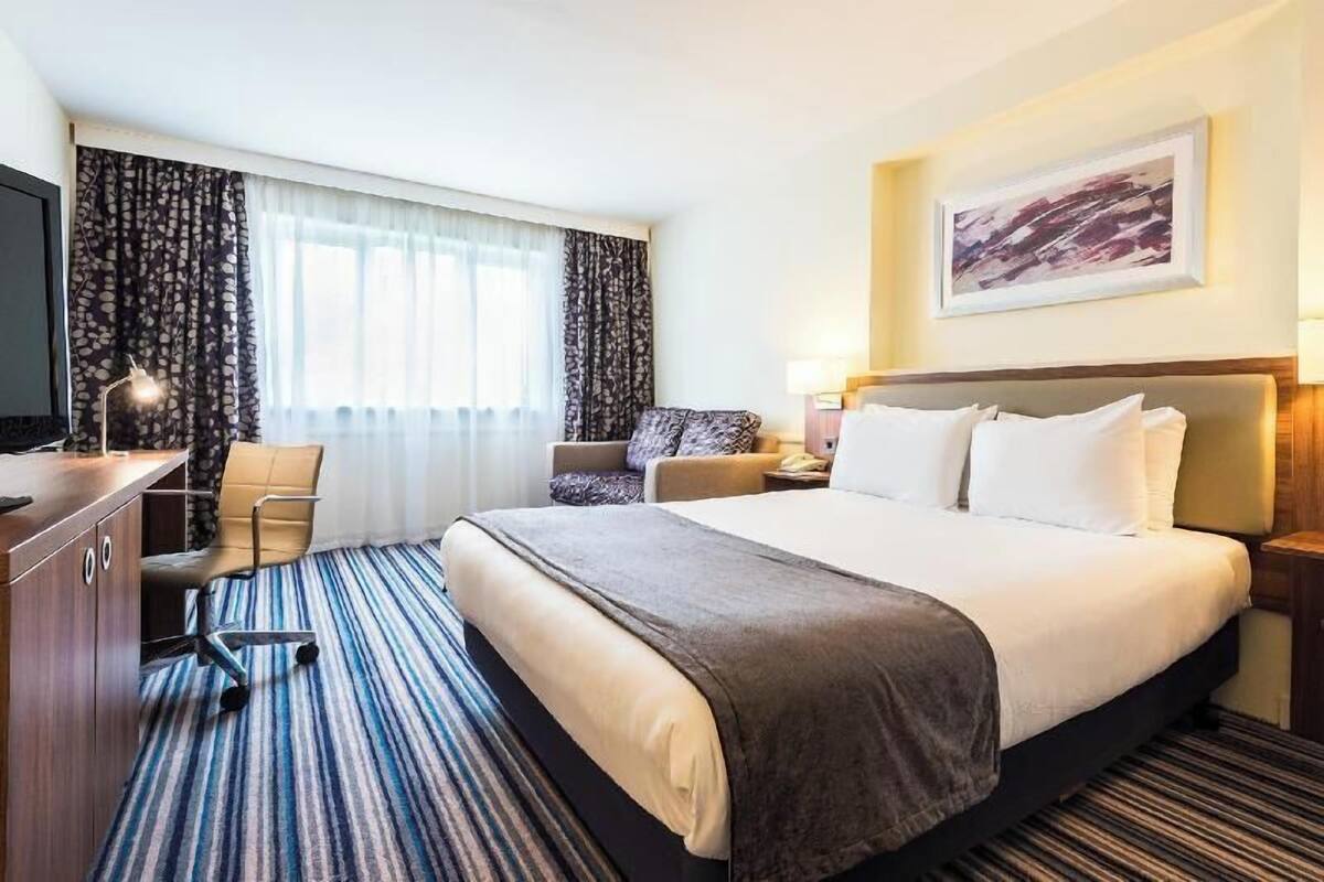 Una habitación de hotel con cama, escritorio, silla, televisor y ventana con cortinas.