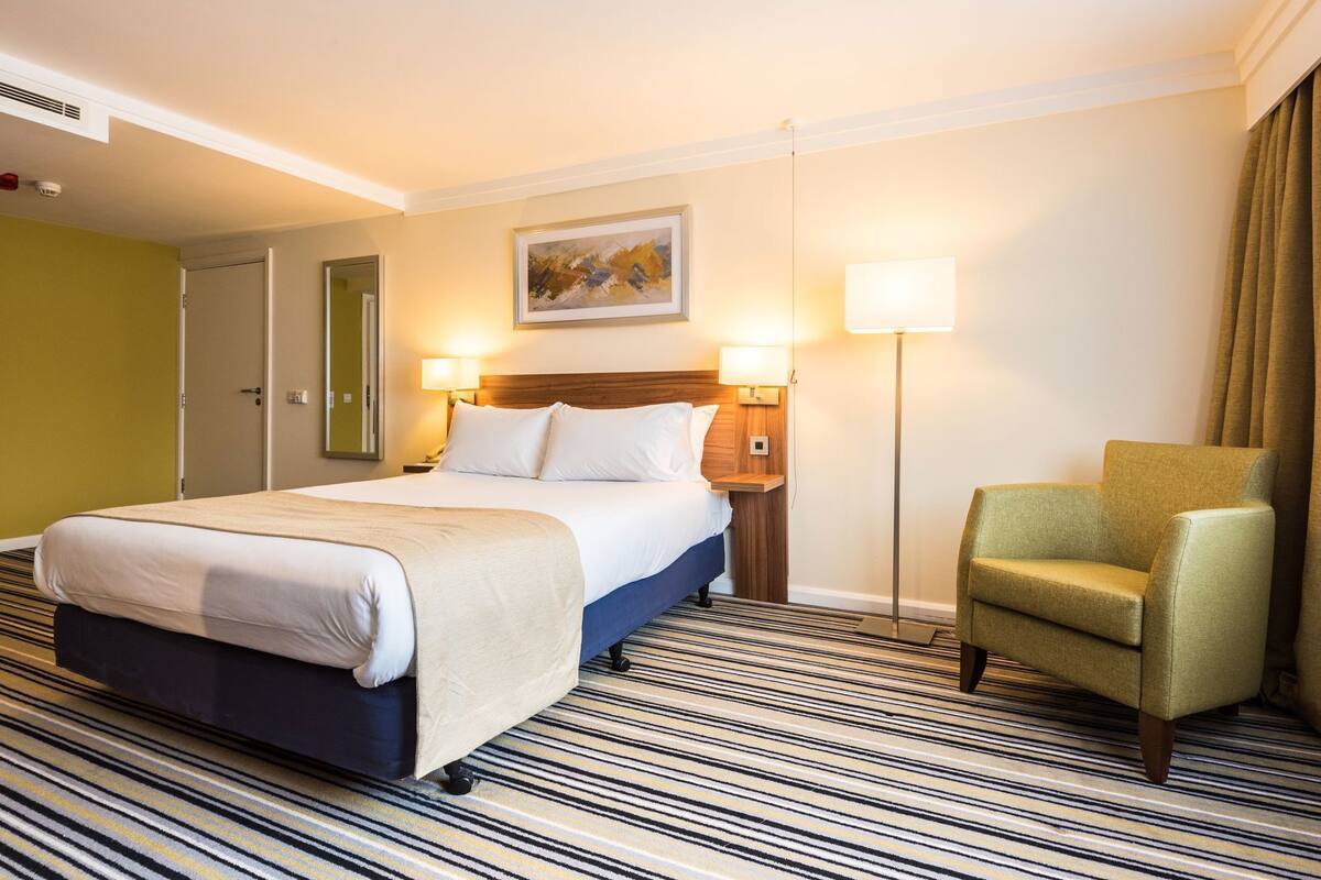 Una habitación de hotel con cama, lámparas de noche, una silla y un cuadro en la pared.