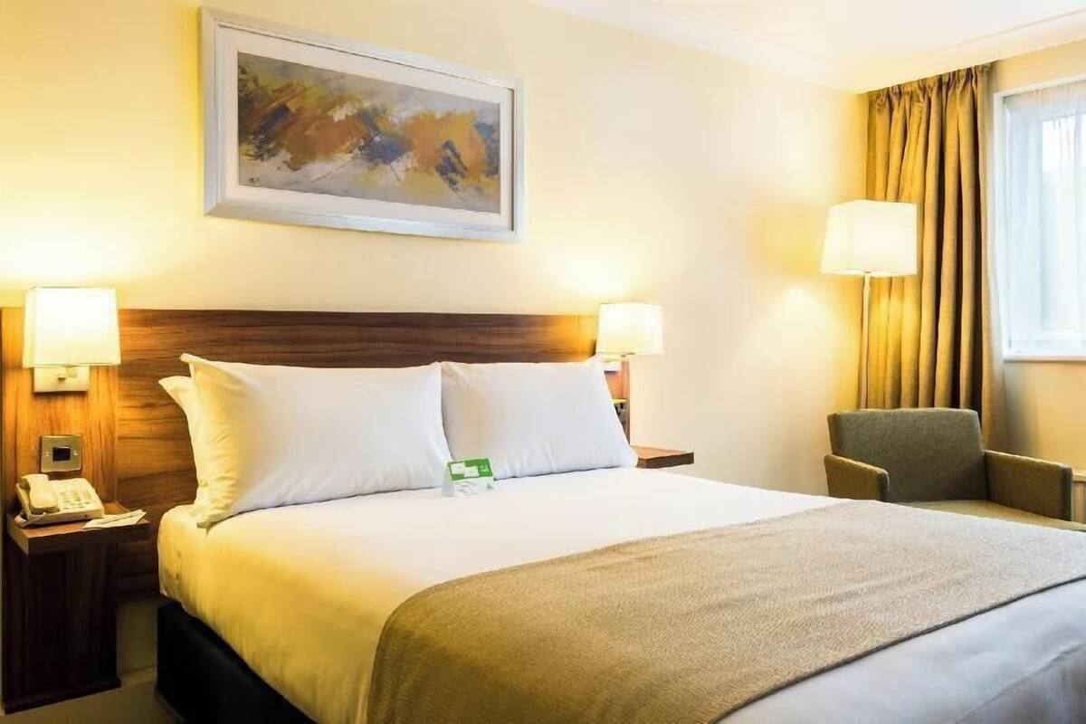 Habitación de hotel con una cama grande, dos lámparas de noche, una silla, un cuadro en la pared y una ventana con cortinas.