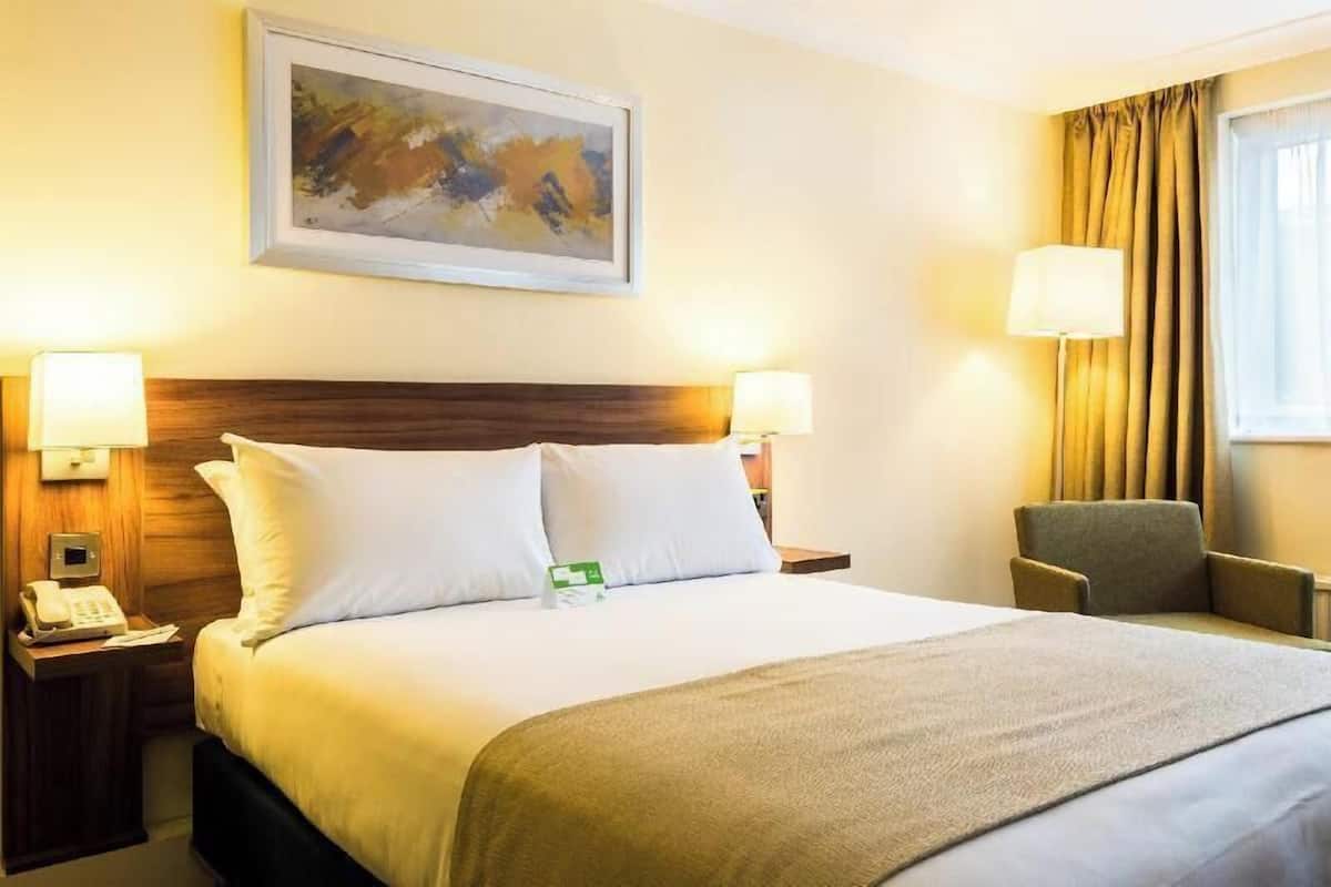 Hotellihuone, jossa on suuri sänky, kaksi yöpöytävalaisinta, tuoli, taulu seinällä ja ikkuna, jossa on verhot.