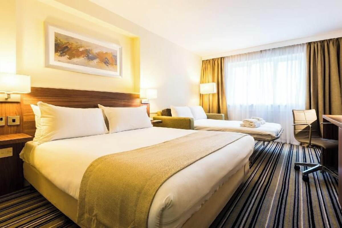A hotel room with two beds, a desk, a chair, a lamp, and a painting on the wall.
