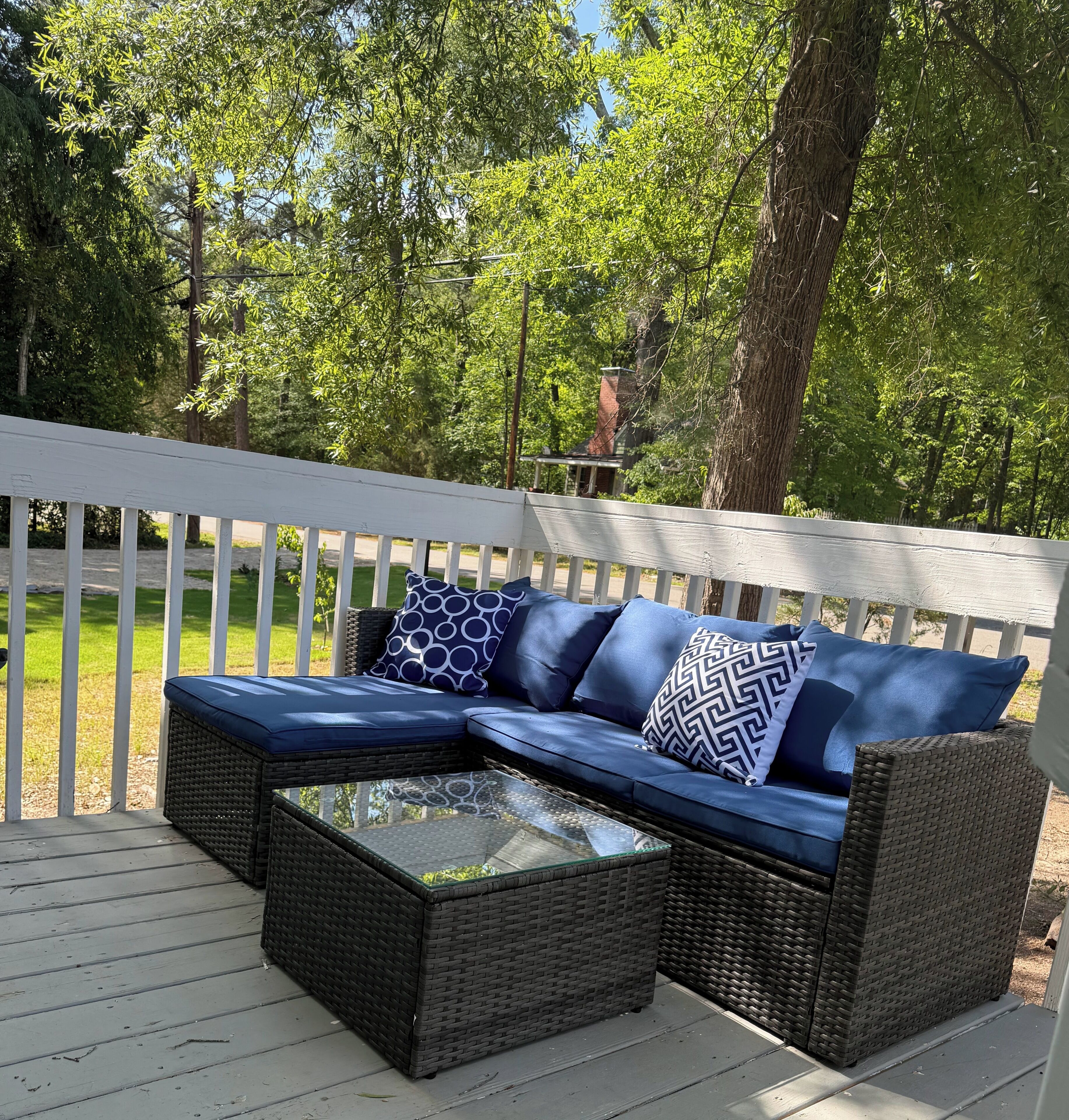 An outdoor seating area with a wicker sofa and a glass-top coffee table, surrounded by a white deck and lush greenery.