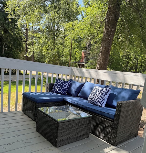 An outdoor seating area with a wicker sofa and a glass-top coffee table, surrounded by a white deck and lush greenery.