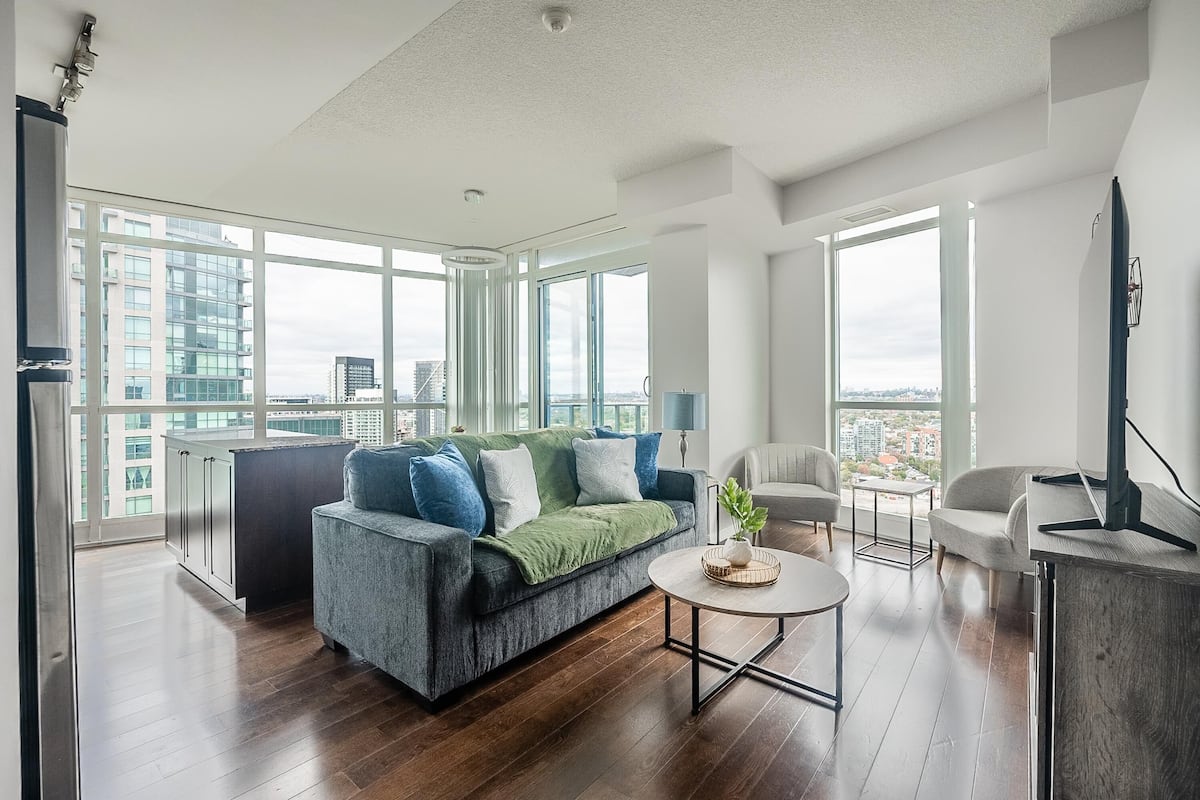 A spacious living room with a grey sofa, a coffee table, and a TV. Large windows offer a view of the cityscape.