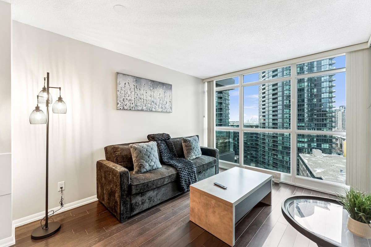 A modern living room with a sofa, a coffee table, a floor lamp, and a large window with a city view.