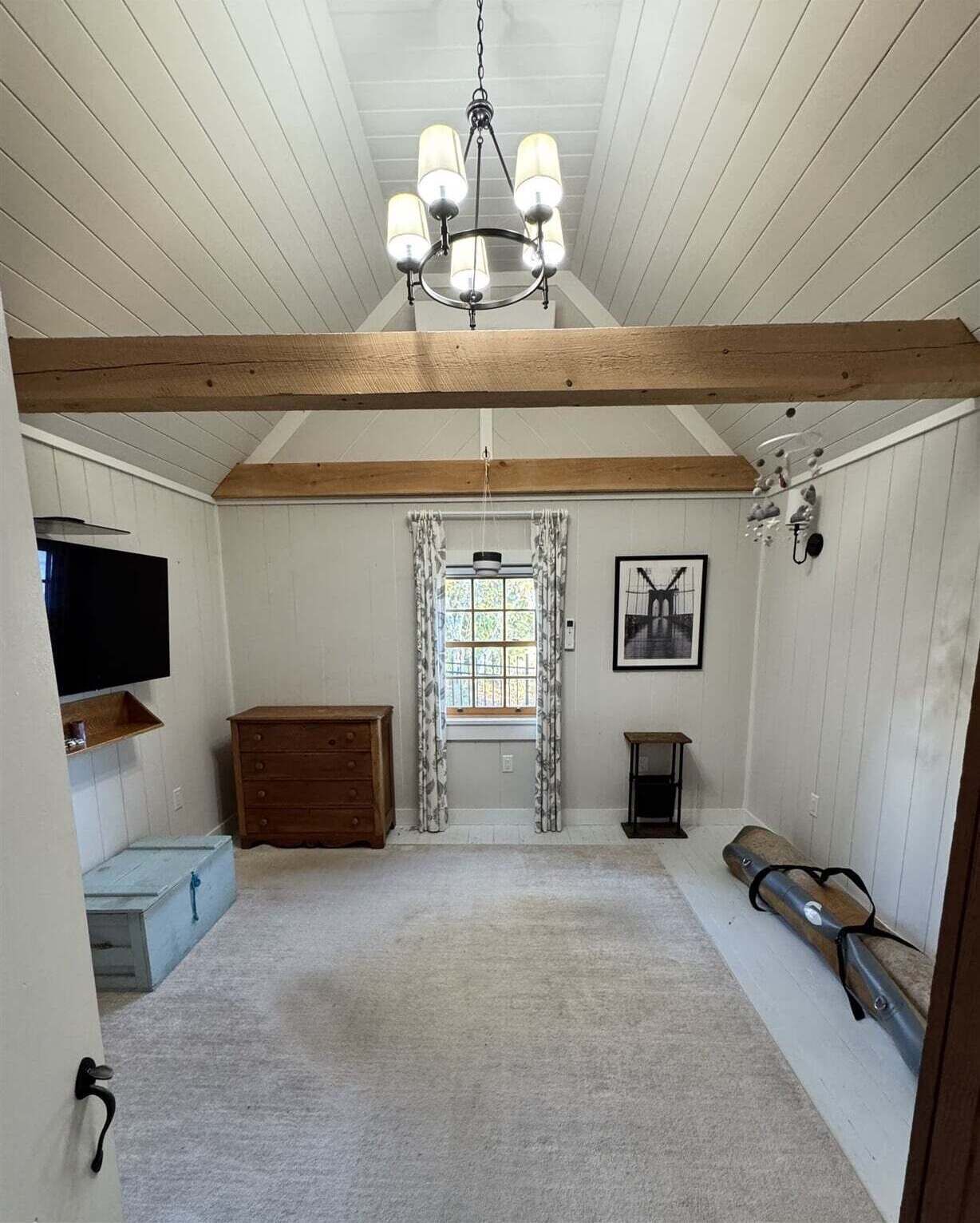 A room with a wooden ceiling, a chandelier, a window with curtains, a television, a dresser, a framed picture, and a bench.