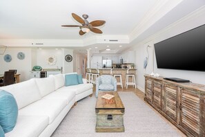 Uma sala de estar com sofá branco, mesa de centro, ventilador de teto, área de cozinha e TV fixada na parede.