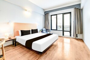 Une chambre d’hôtel avec un lit, un bureau avec une lampe, une chaise, une fenêtre avec des rideaux et une vue sur les bâtiments environnants.
