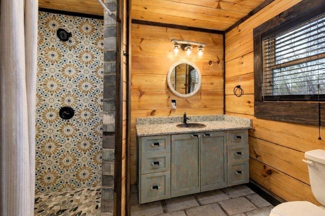 Un baño con detalles de madera, un lavamanos con encimera de granito, un espejo y una ventana con persianas.