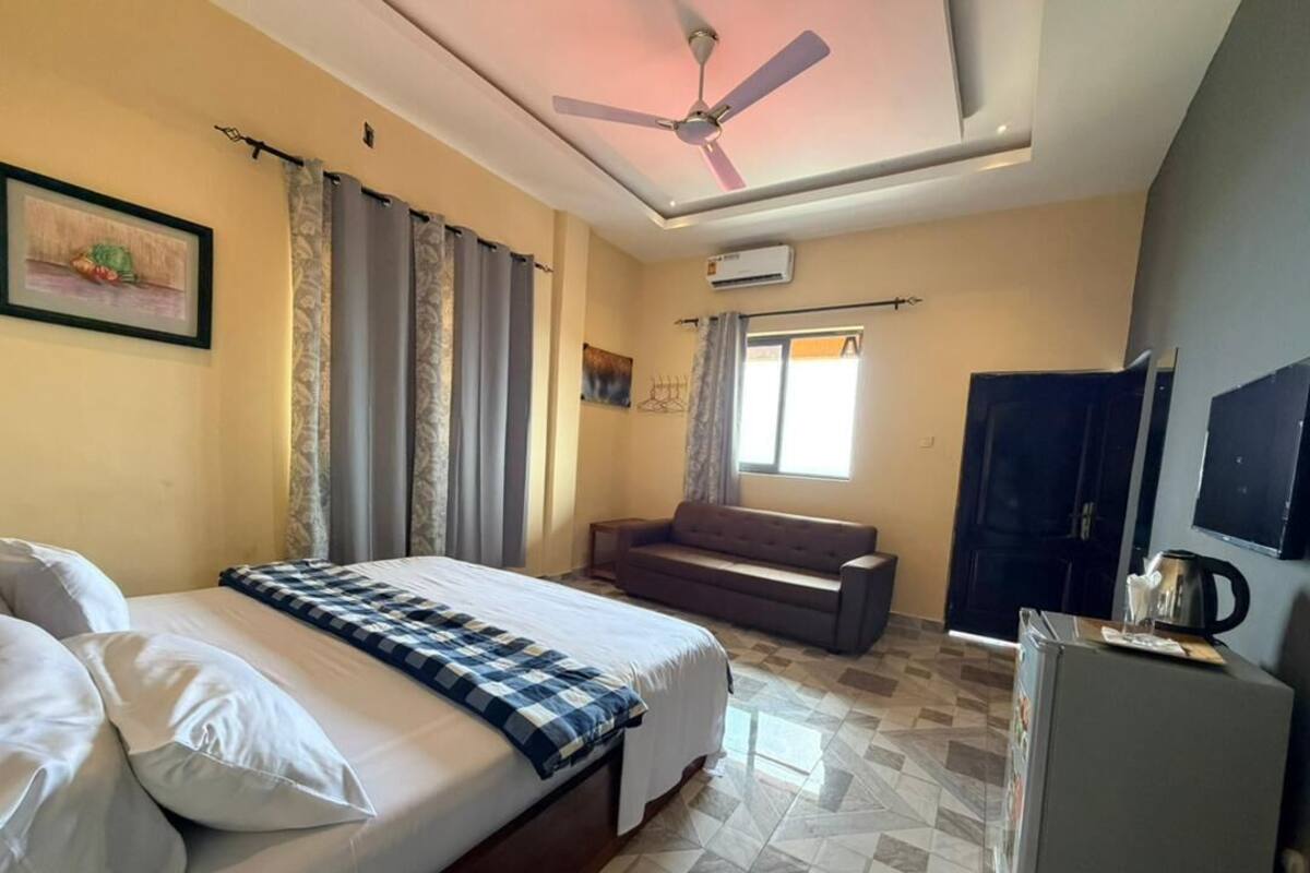 A hotel room with a bed, a sofa, a ceiling fan, an air conditioning unit, a window with curtains, a television, and a refrigerator.