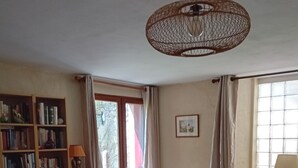 Ein Wohnzimmer mit Bücherregal, einem Korbsessel, einem Tisch mit Tablett, einer Topfpflanze und einem Fenster mit Vorhängen.