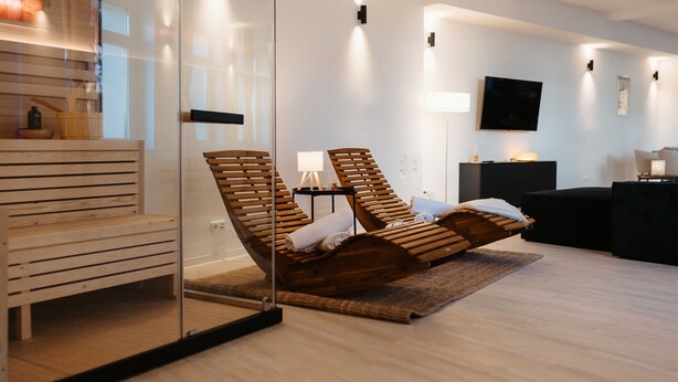 A modern room with a glass partition, wooden lounge chairs, a wooden bench, a black sofa, and a flat-screen TV mounted on the wall.