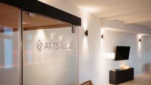 A modern interior with a glass wall displaying the logo 'ARTSAUNA,' a flat-screen TV mounted on the wall, and a minimalist furniture arrangement.