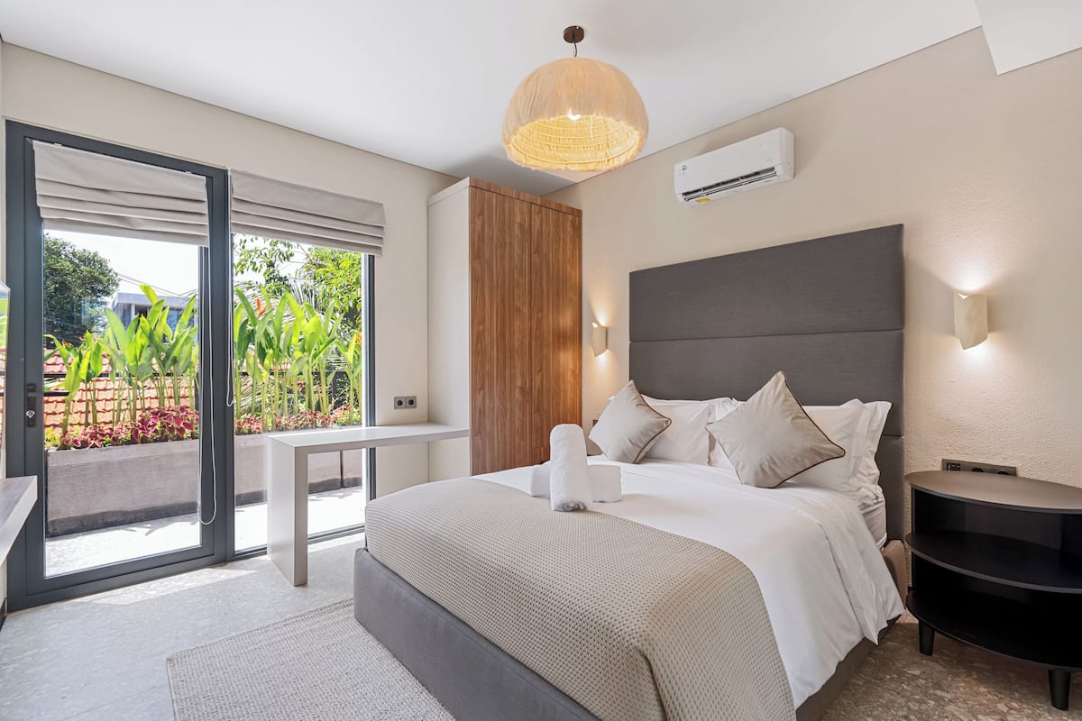 A modern bedroom with a bed, bedside table, and a view of greenery outside.