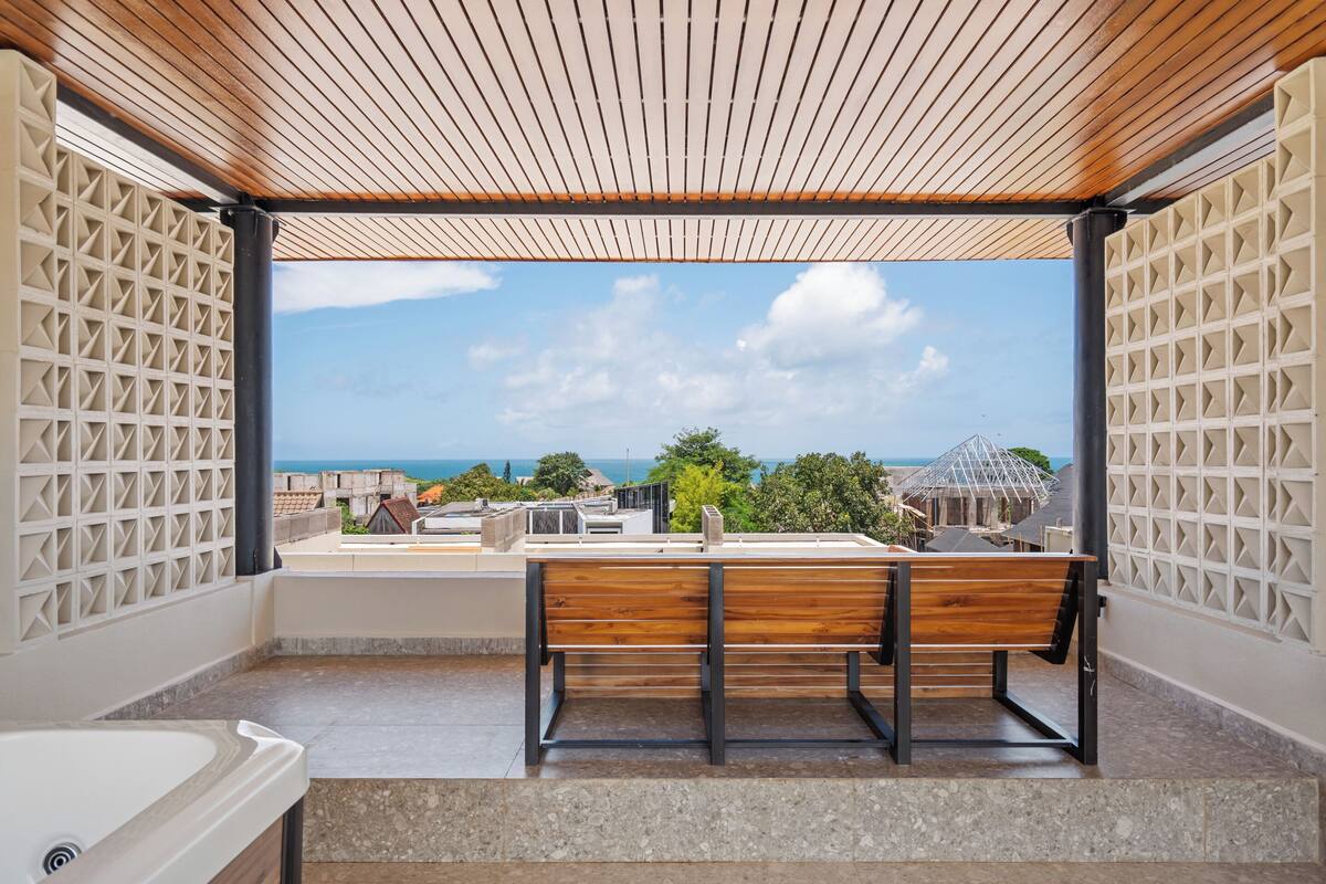 A balcony with a wooden bench, a view of the sea, and a white bathtub.