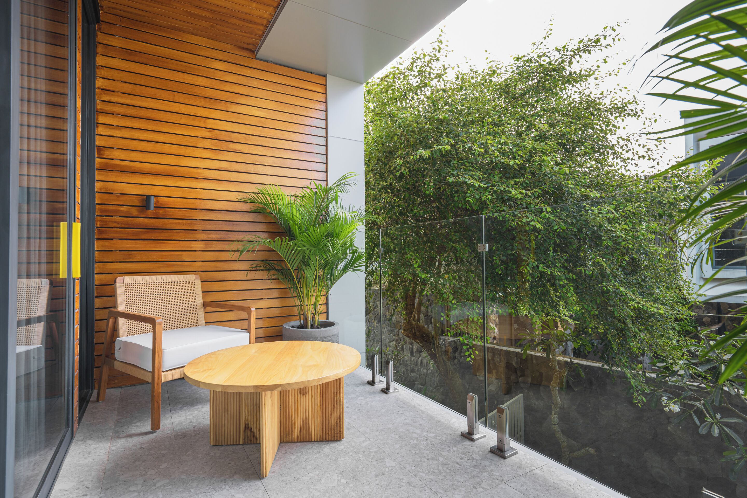 A modern outdoor seating area with a wooden table and chairs, a potted plant, and a glass railing overlooking a lush garden.