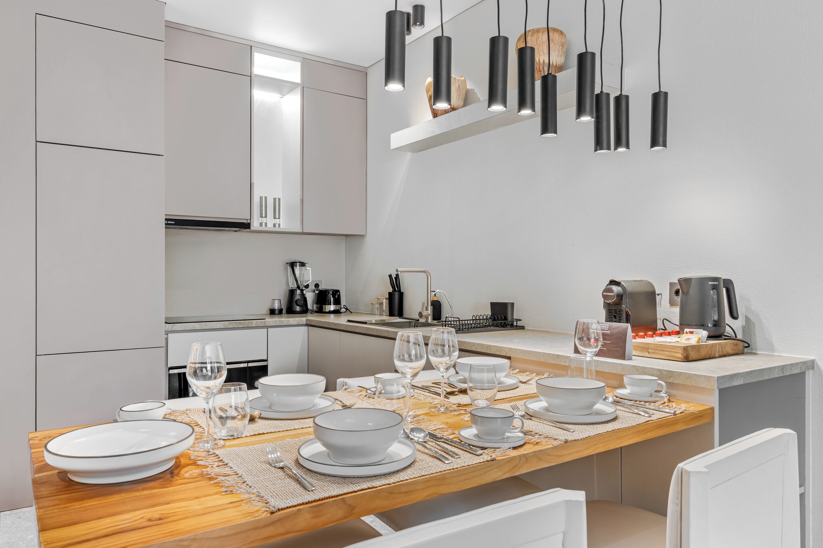 Uma cozinha moderna com armários brancos, uma mesa de jantar de madeira posta com pratos e copos brancos, e iluminação pendente.