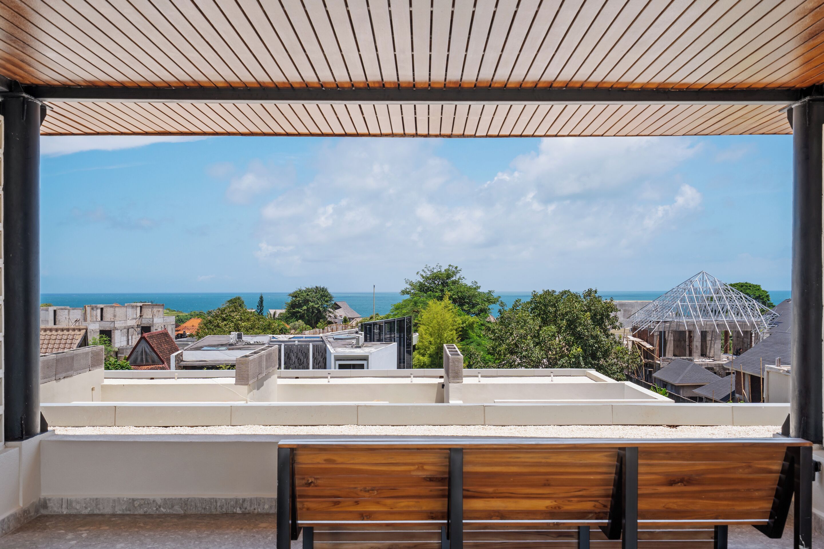 A balcony with a wooden bench, a view of the sea, and buildings in the background.