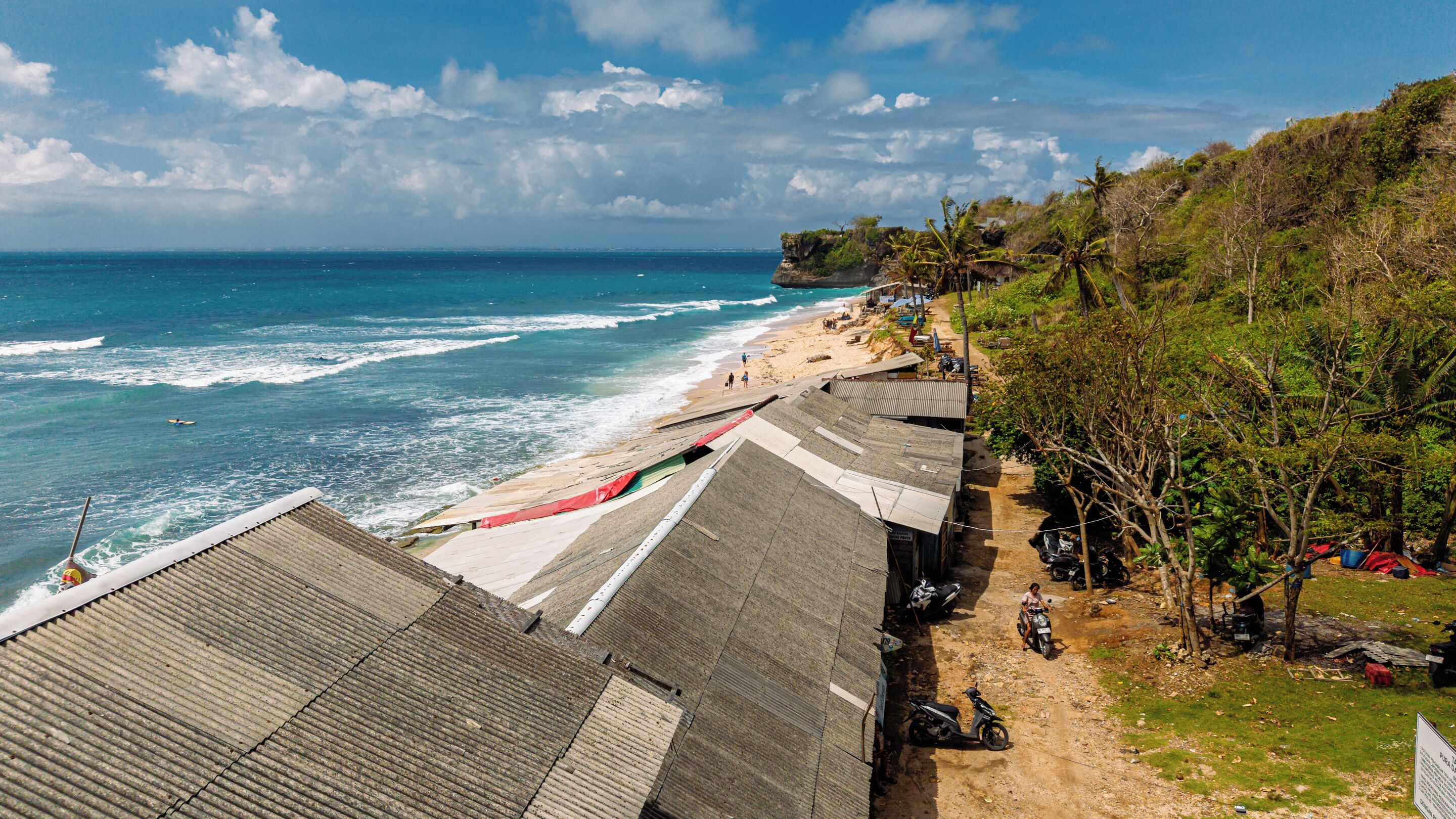 A coastal view with a beach, ocean waves, and a dirt path leading to a row of buildings.