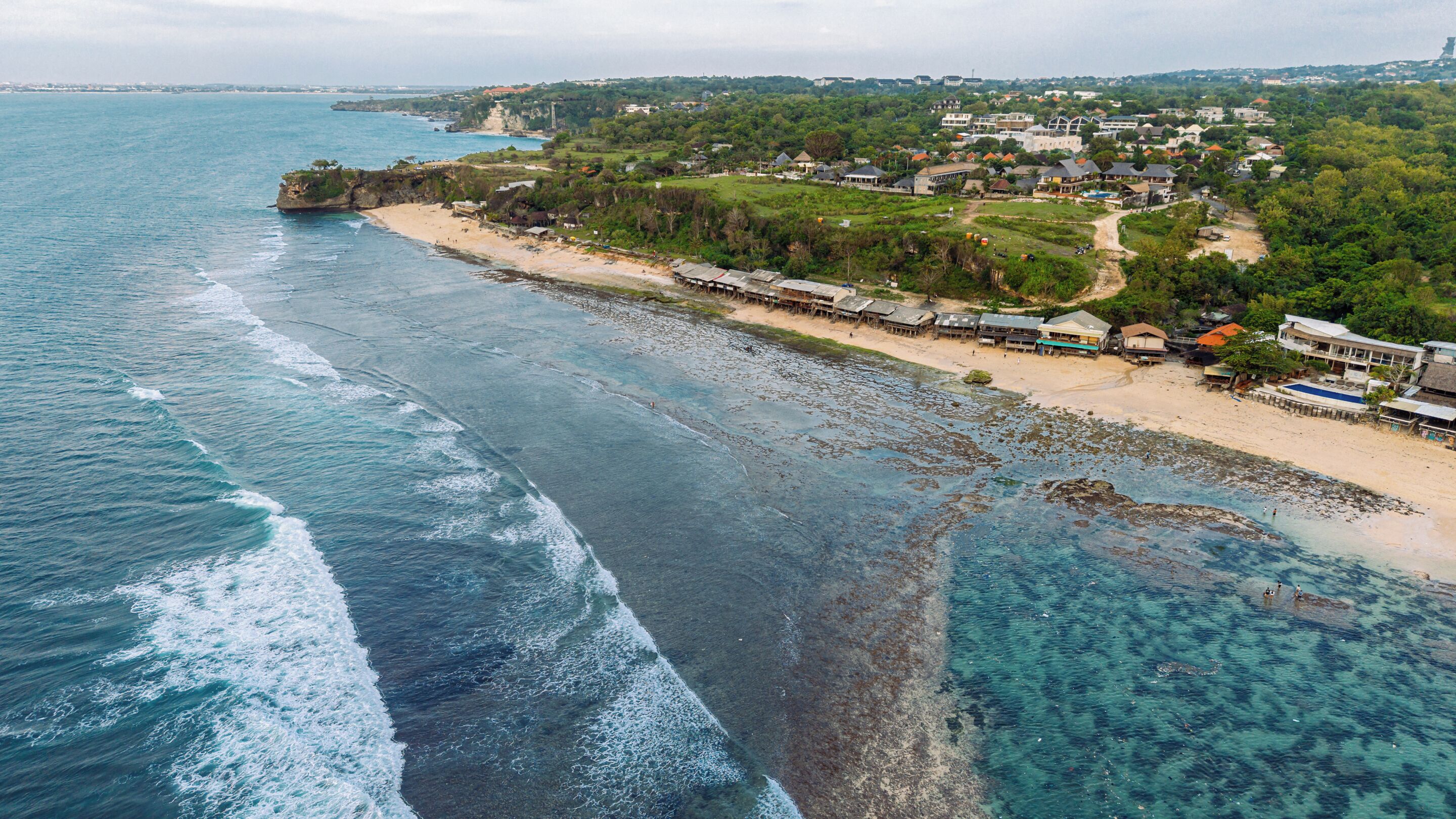 An aerial view of a coastal area with a beach, clear waters, and a residential area with houses and a pier.