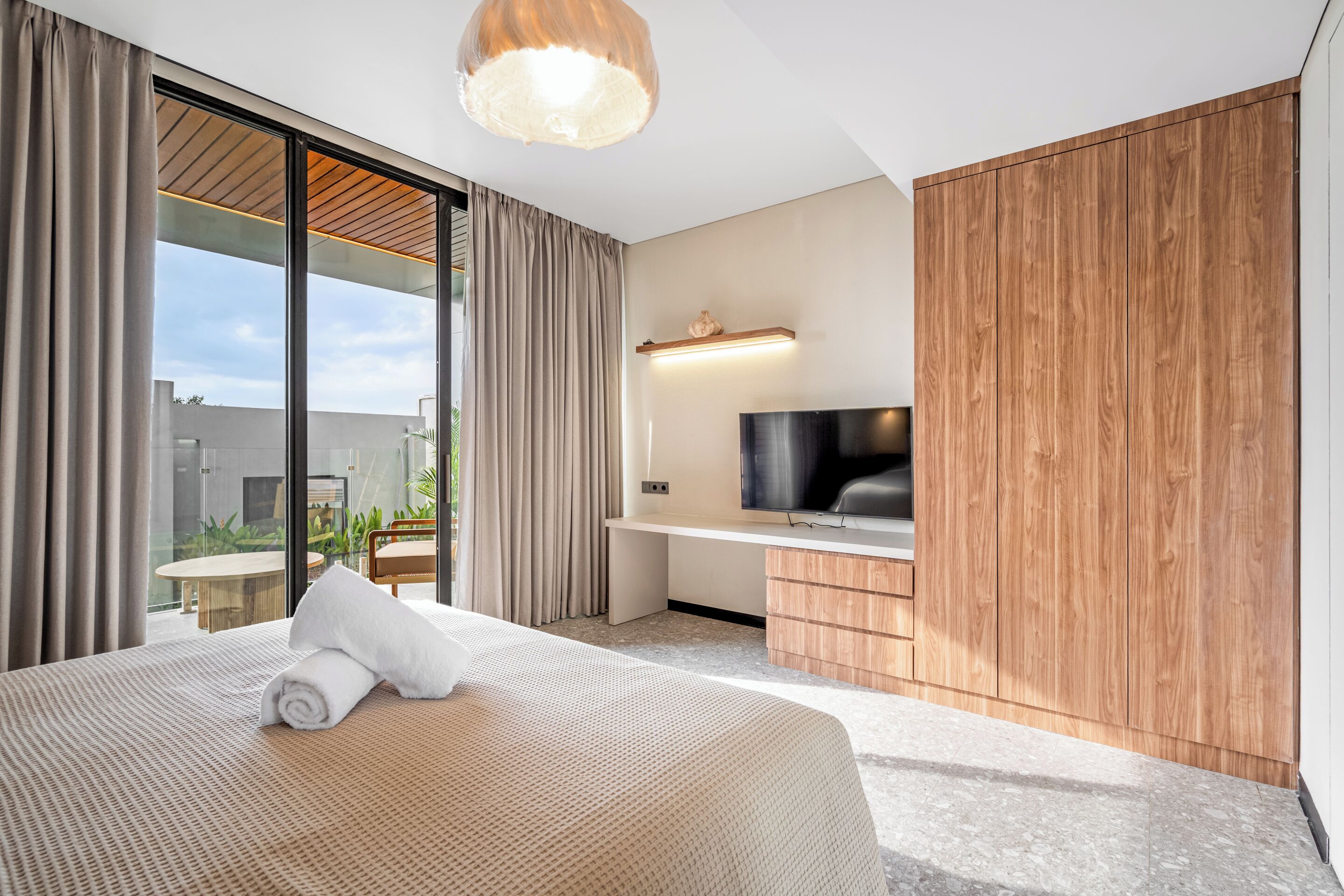 A modern hotel room with a large bed, a flat-screen TV, a wooden wardrobe, and a sliding glass door to the outside.