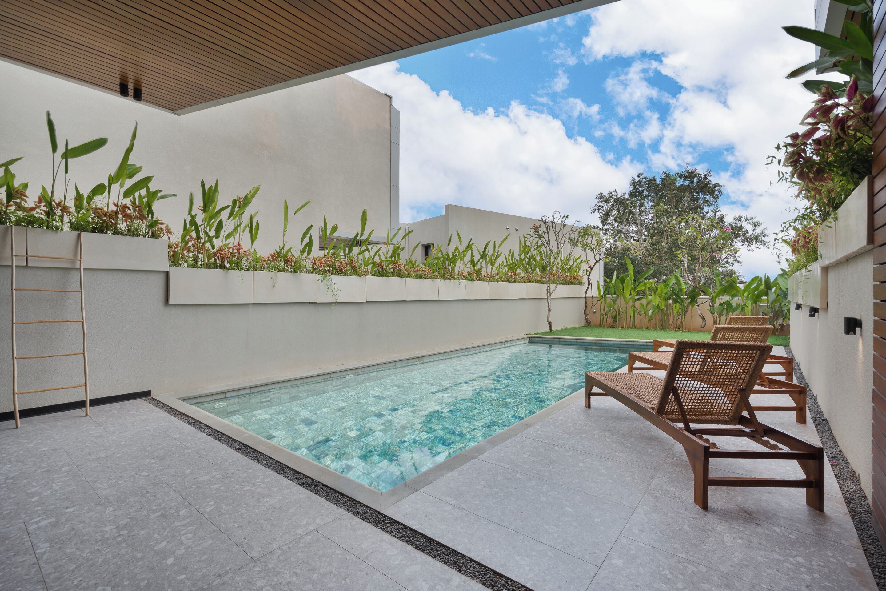 A swimming pool with lounge chairs and a wooden ladder, surrounded by greenery and a clear sky.