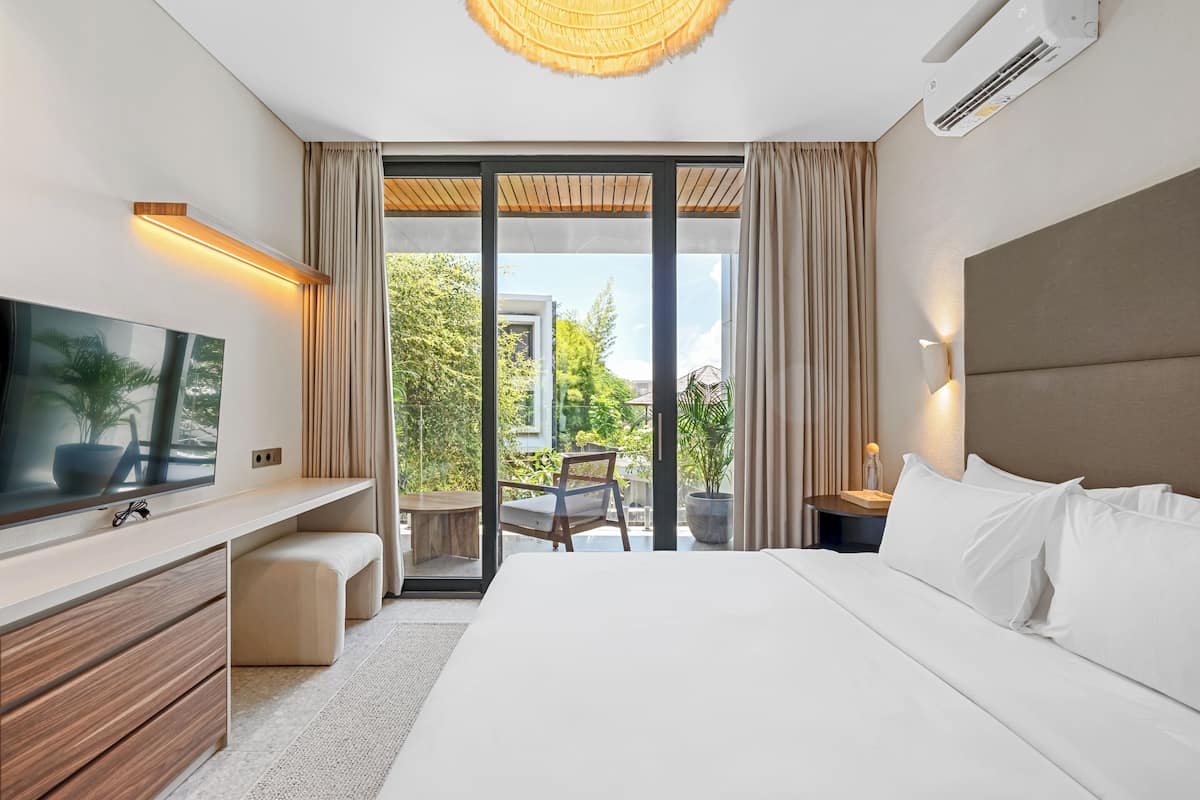 A modern hotel room with a large bed, a flat-screen TV, a desk, and a balcony with a view of greenery.