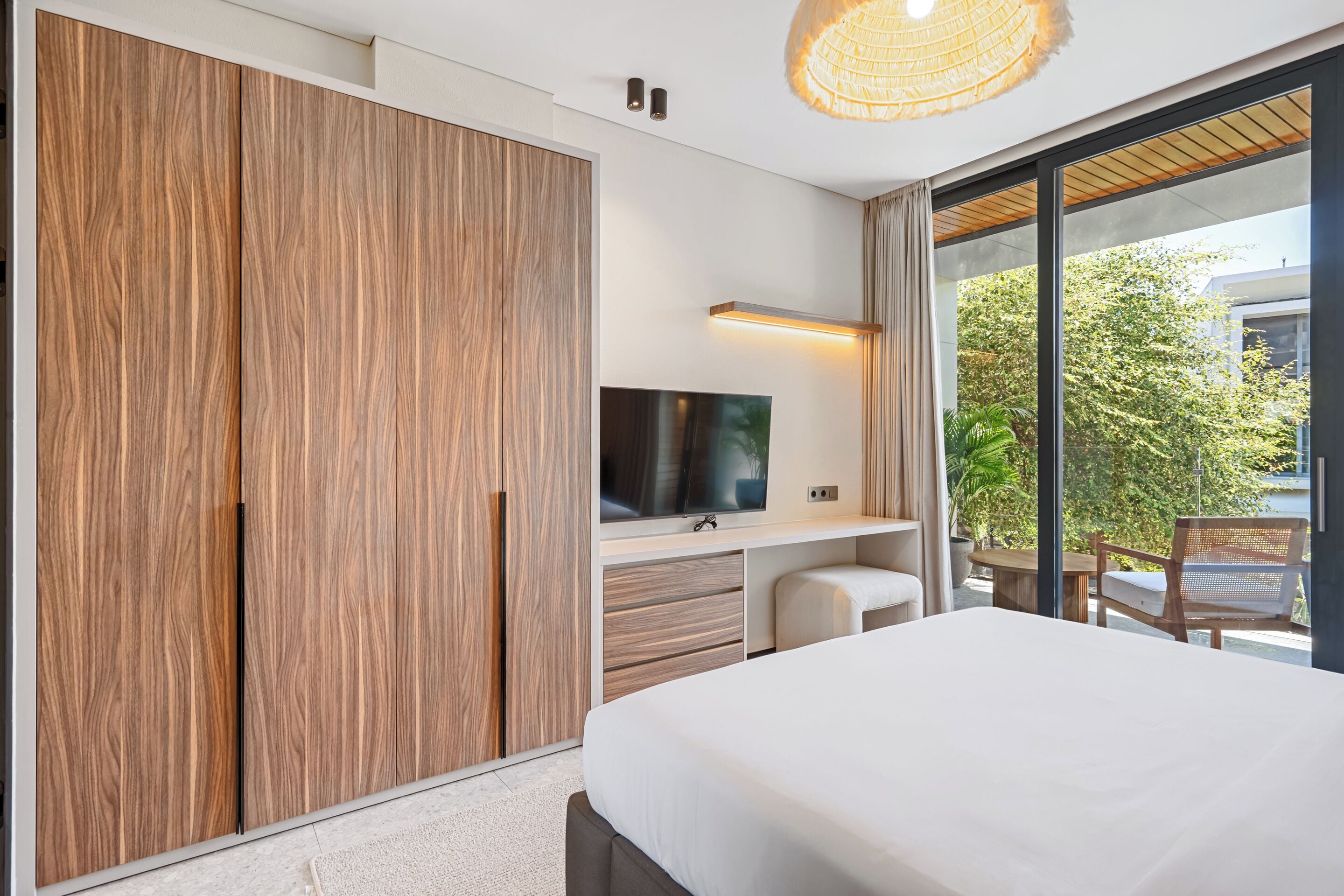 A modern bedroom with a large bed, a TV, a wardrobe, and a balcony with a view of trees.