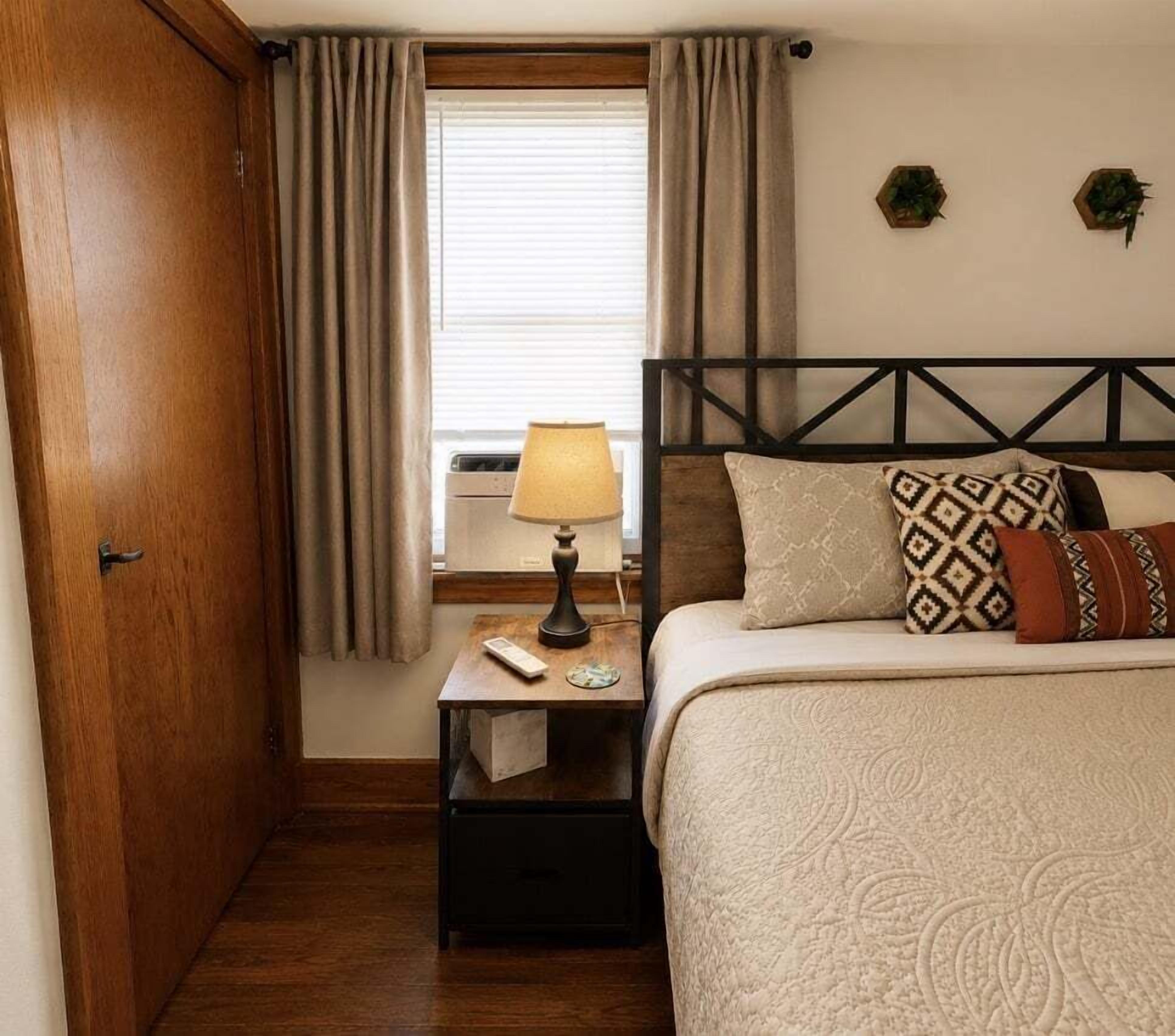 A bedroom with a bed, a nightstand, a lamp, a window with curtains, and a door.