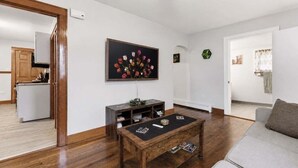 A living room with a wooden coffee table, a grey sofa, a TV on the wall, and a kitchen in the background. - Johnstown Stay Near Laurel Highlands & Hiking (Johnstown)