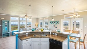A kitchen with a central island, a dining area with a table and chairs, and a view through large windows. - Toes In The Sand 3 Bedroom Home (Port Bolivar)